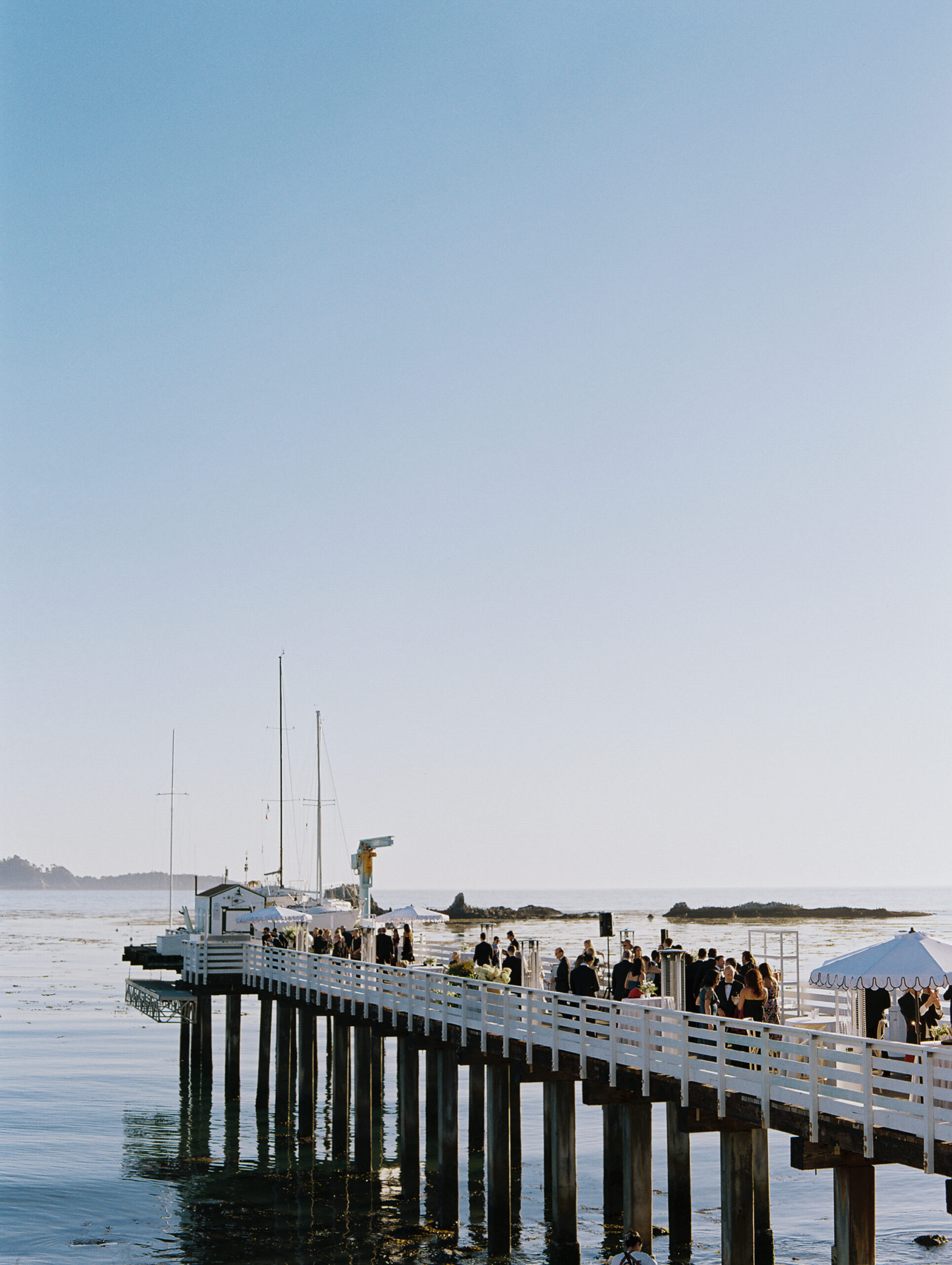 Pebble beach and tennis club pier holds guests enjoying cocktail hour in the distance at a summer wedding.